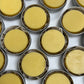 Multiple small round yellow lotion bars in metal tins on a white background

